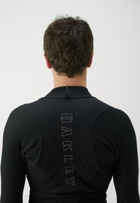 Black athletic long-sleeve top with a textured pattern, featuring an "OAKLEY" logo vertically on the back and smooth fabric texture.