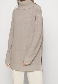 Femme portant un pull à col roulé en grosse maille beige oversize avec des poignets côtelés et des fentes latérales, associé à un pantalon clair.