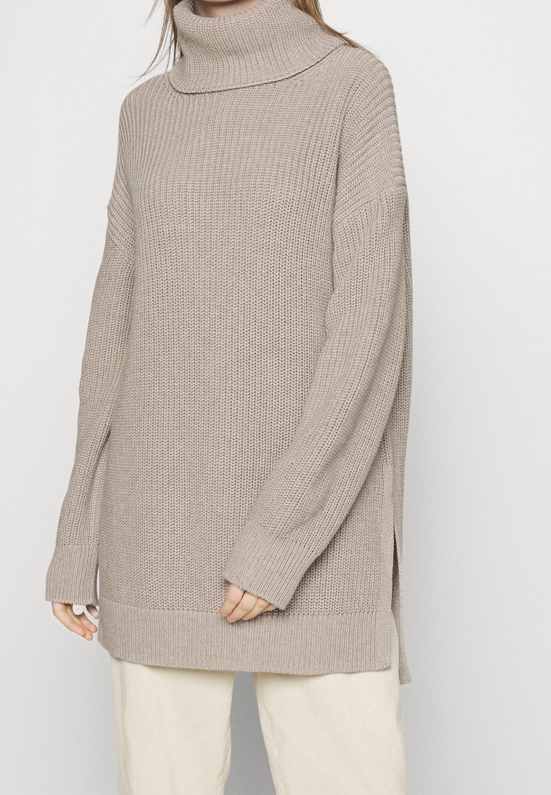 Femme portant un pull à col roulé en grosse maille beige oversize avec des poignets côtelés et des fentes latérales, associé à un pantalon clair.