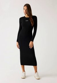 Robe noire côtelée à manches longues avec une silhouette ajustée, longueur au genou, ornée d'un petit logo blanc au niveau du col, associée à des baskets blanches.