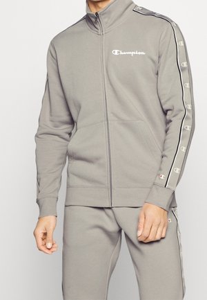 Lysegrå sweatjakke med lynlås i blødt stof. Har høj krave, forlommer og logo på brystet med kontrasterende stribe ned ad ærmerne.