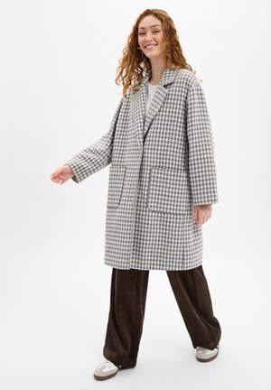 Grijs-wit houndstooth-gepatenteerde jas met uitgesneden revers, twee voorklauwen, een licht oversized pasvorm, gedragen over bruine corduroybroek.