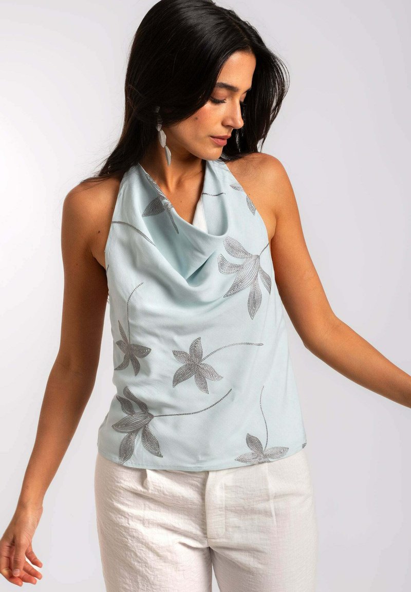 Top sans manches bleu clair, avec un col drapé, arborant des broderies en fleurs argentées et des motifs de feuilles délicats. Associé à un pantalon en lin blanc.