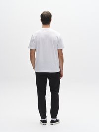 Balta kokvilnas T-krekls ar vaļīgu piegriezumu un īsām piedurknēm. Kombinēts ar melnām sašaurinātām biksēm un melnām sporta kurpēm, stāvot pret vienkāršu baltu fonu.