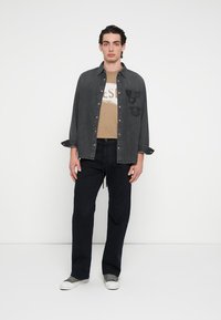 Svart denimskjorte over en tan farget grafisk t-skjorte, kombinert med mørke bukser og grå joggesko. Skjorten har lapper og en avslappet passform.