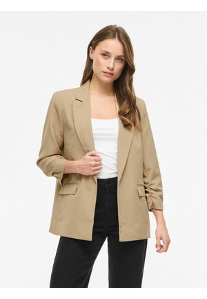 Femme aux longs cheveux bruns portant un blazer beige sur un haut blanc et un pantalon noir, debout devant un fond blanc.