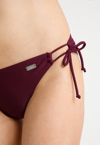 Burgunderfarbene Bikini-Hosen mit verstellbaren Seitenschnüren, glattem Stoff und einem kleinen metallischen Logo-Detail am Bund.