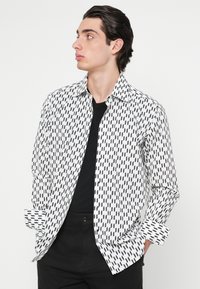 Fehér, fekete geometriai mintás ing, hosszú ujjú és klasszikus gallérral. Fekete póló fölött viselve, sötét nadrággal párosítva.