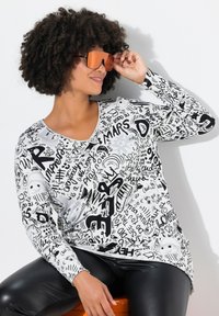 Maglia a maniche lunghe bianca con testo nero e motivi grafici. Presenta un colletto a V e una vestibilità morbida. Il modello indossa occhiali da sole riflettenti arancioni.