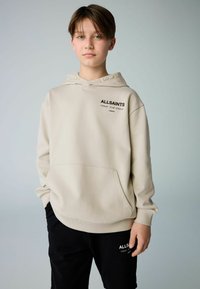 Sudadera beige con capucha y bolsillo canguro, con un logo de texto negro. Lleva puestos pantalones negros que presentan un logo a juego en la pierna.