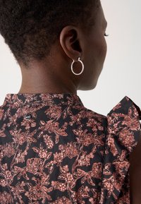 Femme aux cheveux courts et bouclés portant une boucle d'oreille argentée en créole et un haut noir à motif floral rose avec des manches à volants, tournée de dos.