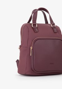 Piccola zainetto borgogna con tasca frontale a granuli, zip dorata, manici doppi corti e logo del marchio discreto sulla tasca.
