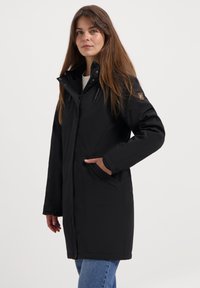 Femme aux longs cheveux bruns portant un manteau d'hiver noir à capuche et un jean bleu, avec une main dans la poche du manteau, se tenant contre un fond blanc.