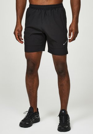 Mand iført sorte atletikshorts og sorte løbesko står mod en ensartet lys baggrund.