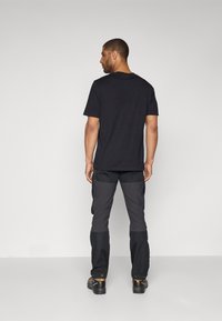 Czarny t-shirt z krótkim rękawem i szare spodnie cargo z łatami na kolanach. Strój uzupełniają solidne czarne obuwie. Gładkie tło.
