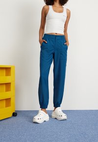 Modré kalhoty s elastickým pasem a kapsami, s elastickými manžetami, vyrobené z hladké látky. Spárovány s bílými robustními dřeváky a bílým tílkem.