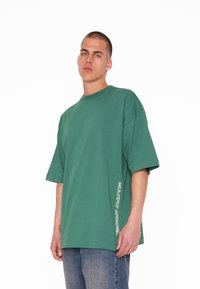 T-shirt verde oversize realizzato in cotone morbido, con collo rotondo e maniche corte. Logo bianco ben visibile sul lato. Sotto, sono indossati jeans di denim.