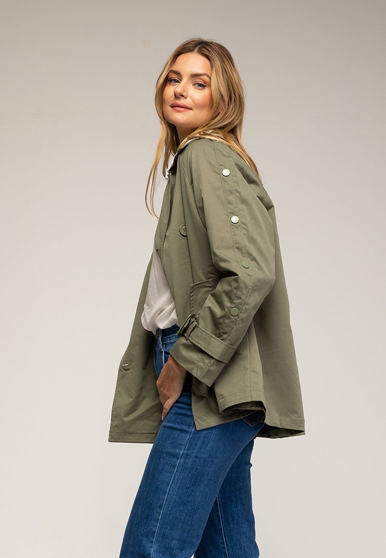 Olivgrüne Jacke mit lockerer Passform, mit Knopfakzenten an den Ärmel, seitlichen Taschen und einem Knopfverschluss, getragen über einem weißen Oberteil.