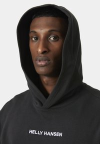 Black hoodie with a large hood, featuring the white text "HELLY HANSEN" embroidered on the front, smooth texture, and casual fit.