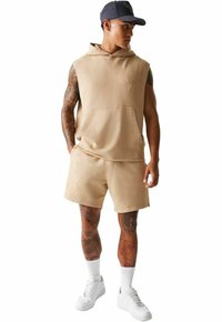 Beige ärmellose Kapuzenjacke mit einer Vorderseite-Tasche, passenden Shorts und weißen Sneakers. Der Stoff hat eine glatte Textur; dezentes Branding-Detail.