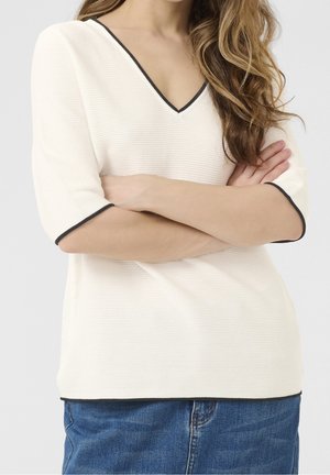 Femme aux longs cheveux ondulés portant un haut blanc à col en V avec une bordure noire et une jupe en jean bleue, debout les bras croisés.