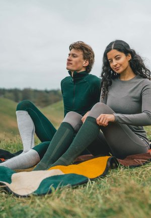 Grüne und graue, eng anliegende Langarm-Shirts und knielange Socken mit strukturierten Mustern, sitzen auf einer bunten, gemusterten Decke auf dem Gras.