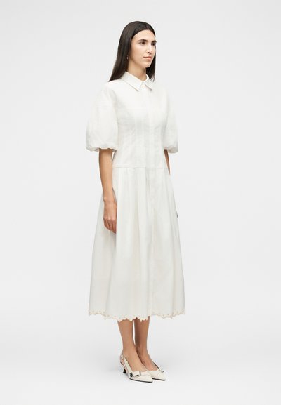 Γυναίκα με μακριά σκούρα μαλλιά φορώντας λευκό μίντι φόρεμα με φουσκωτά μανίκια και κυματιστό τελείωμα, σε συνδυασμό με λευκές γόβες τύπου slingback.