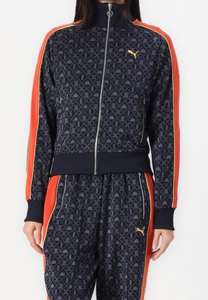 Personne portant un survêtement Puma bleu marine avec un motif géométrique et des bandes latérales rouges et jaunes, debout devant un fond blanc uni.