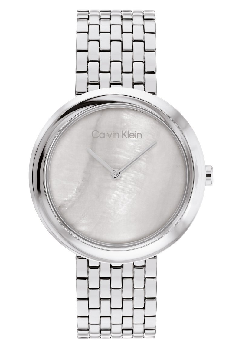 Montre-bracelet en métal argenté avec un boîtier rond, un cadran en nacre blanche, de fines aiguilles argentées, et un bracelet en acier inoxydable tressé.