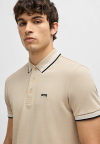 Beige polo-skjorte laget av teksturert stoff, med skjortejakke i svart og hvitt og en liten brodert "BOSS"-logo på brystet.