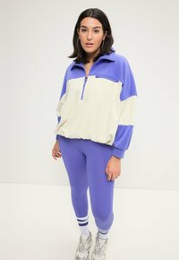 Fleece-Pullover in Creme und Helllila, mit einem Reißverschlusskragen, überschnittenen Schultern und elastischem Saum; kombiniert mit passenden lilafarbenen Leggings.
