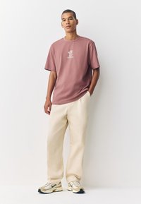 Pink short-sleeve t-shirt with front logo, paired with cream-colored trousers and beige sneakers with green accents. Smooth cotton texture.