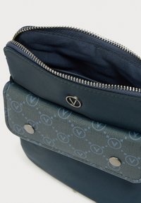 Mala tamnoplava torbica s uzorkom na preklopu, s metalnim drukerima i djelomično otvorenim srebrnim patentnim zatvaračem na vrhu.