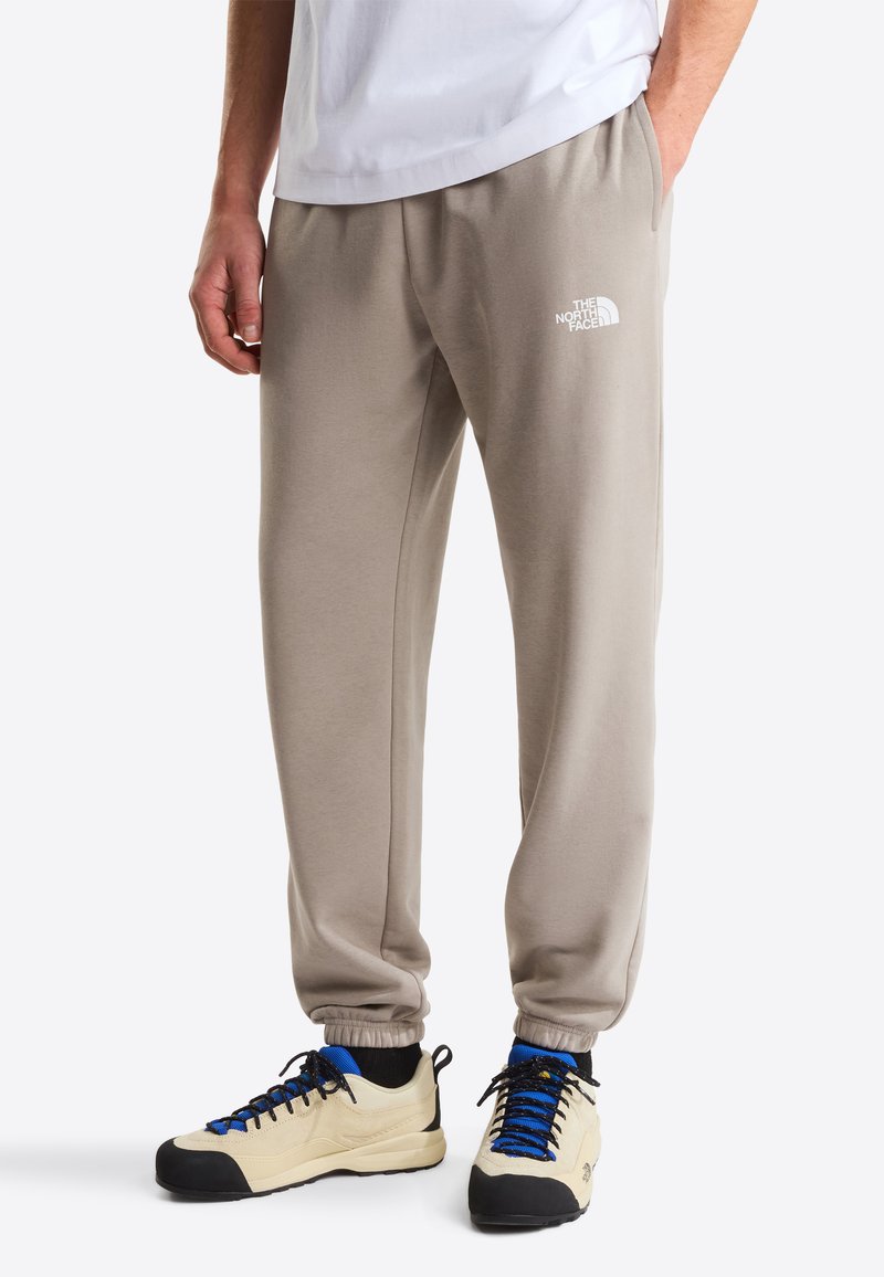 Pantalones de chándal beige con cintura y puños elásticos; presenta un logo blanco en el muslo izquierdo; combinados con zapatillas beige y negras.