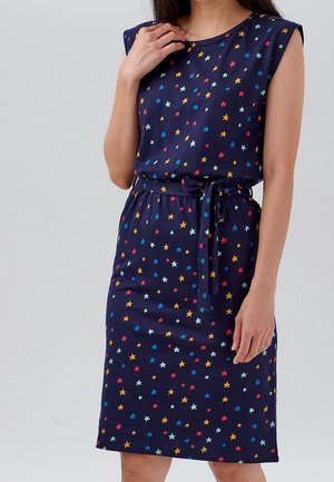 Femme portant une robe bleu marine sans manches avec un motif étoile multicolore et une ceinture nouée à la taille, debout devant un fond uni.