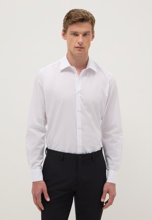 Jonge man die een nauwsluitend wit overhemd draagt, ingestopt in zwarte nette broek, staand tegen een effen lichte achtergrond.