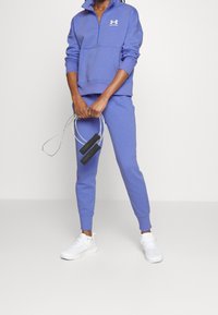 Suéter de forro polar morado con cuello con cremallera, combinado con pantalones jogger a juego. El modelo sostiene una cuerda para saltar y lleva zapatillas deportivas blancas.