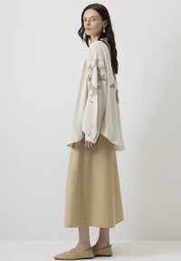 Tunică oversized bej cu broderie complexă pe mâneci, asortată cu o fustă bej și pantofi tip slip-on. Material neted și croială lejeră.