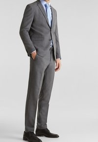 Completo grigio su misura realizzato in tessuto texturizzato, caratterizzato da una giacca monopetto e pantaloni dritti, abbinato a una camicia azzurro chiaro e scarpe nere.