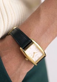 Montre rectangulaire dorée avec un cadran blanc, bracelet en cuir noir et index d'heure discrets. Portée au poignet, mettant en valeur l'élégance.