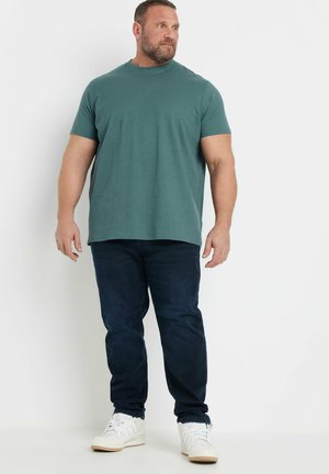 Homme musclé debout, portant un t-shirt bleu sarcelle à manches courtes, un jean foncé et des baskets blanches, regardant légèrement vers la droite.