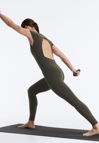 Woman in olive green open-back workout suit stretches on black mat, holding a small dumbbell in one hand.