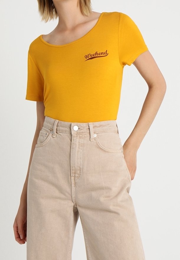 Femme portant un haut jaune moutarde à manches courtes avec le texte « Weekend » et un pantalon beige taille haute, debout avec une main dans la poche.