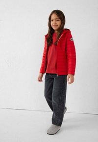 Rode gewatteerde jas met capuchon, gladde textuur en logo op de mouw. Gedragen over een rood shirt, gecombineerd met donkergrijze spijkerbroek en grijze pantoffels.