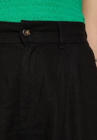 Gros plan sur un pantalon en lin noir avec fermeture à bouton et un haut texturé vert rentré, montrant les détails de la taille et des poches.