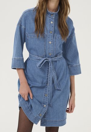 Kvinde iført blå denim knap-op kjole med forlommer og bundet bælte i taljen, poserer med hånd, der holder kanten.