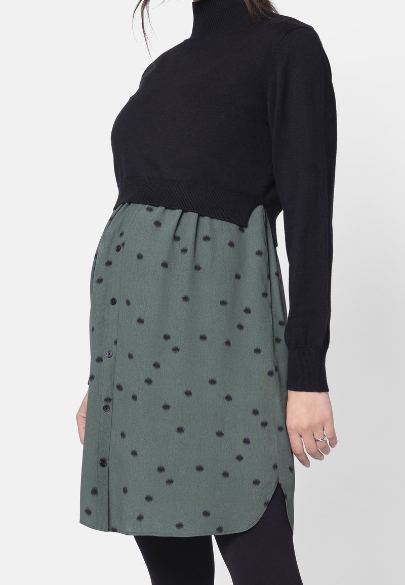 Sukienka ciążowa z czarnym dzianinowym topem oraz zieloną spódnicą z materiału w czarne kropki; detale z guzikami wzdłuż szwu bocznego.