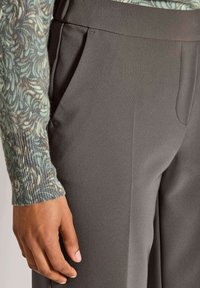 Hand rustend naast donkergrijze, op maat gemaakte broek met zijzak, gecombineerd met een groen gemusterd shirt met lange mouwen.