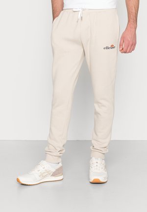 Pantalon de jogging beige avec taille à cordon de serrage et logo ellesse sur la cuisse gauche, porté avec des baskets blanches et beiges sur un sol gris.