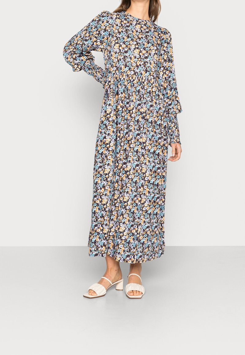 Femme portant une robe fleurie à manches longues, longueur cheville, avec des fleurs bleues et jaunes, associée à des sandales blanches à talons.